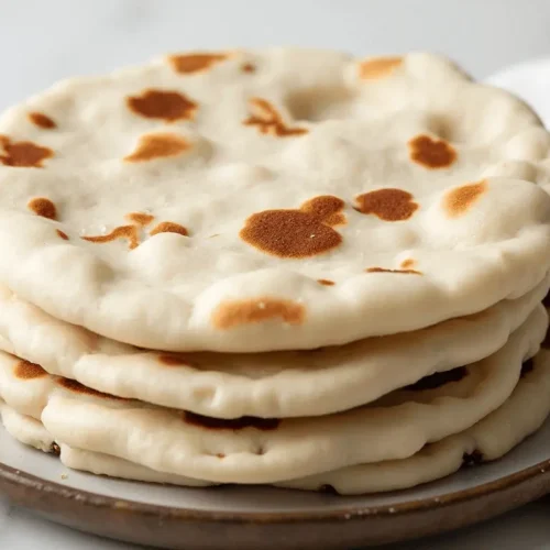 No-Waste Baking: How to Make Sourdough Discard Naan 8 A stack of freshly cooked flatbreads with golden brown spots, served on a plate, accompanied by a folded kitchen towel in the background